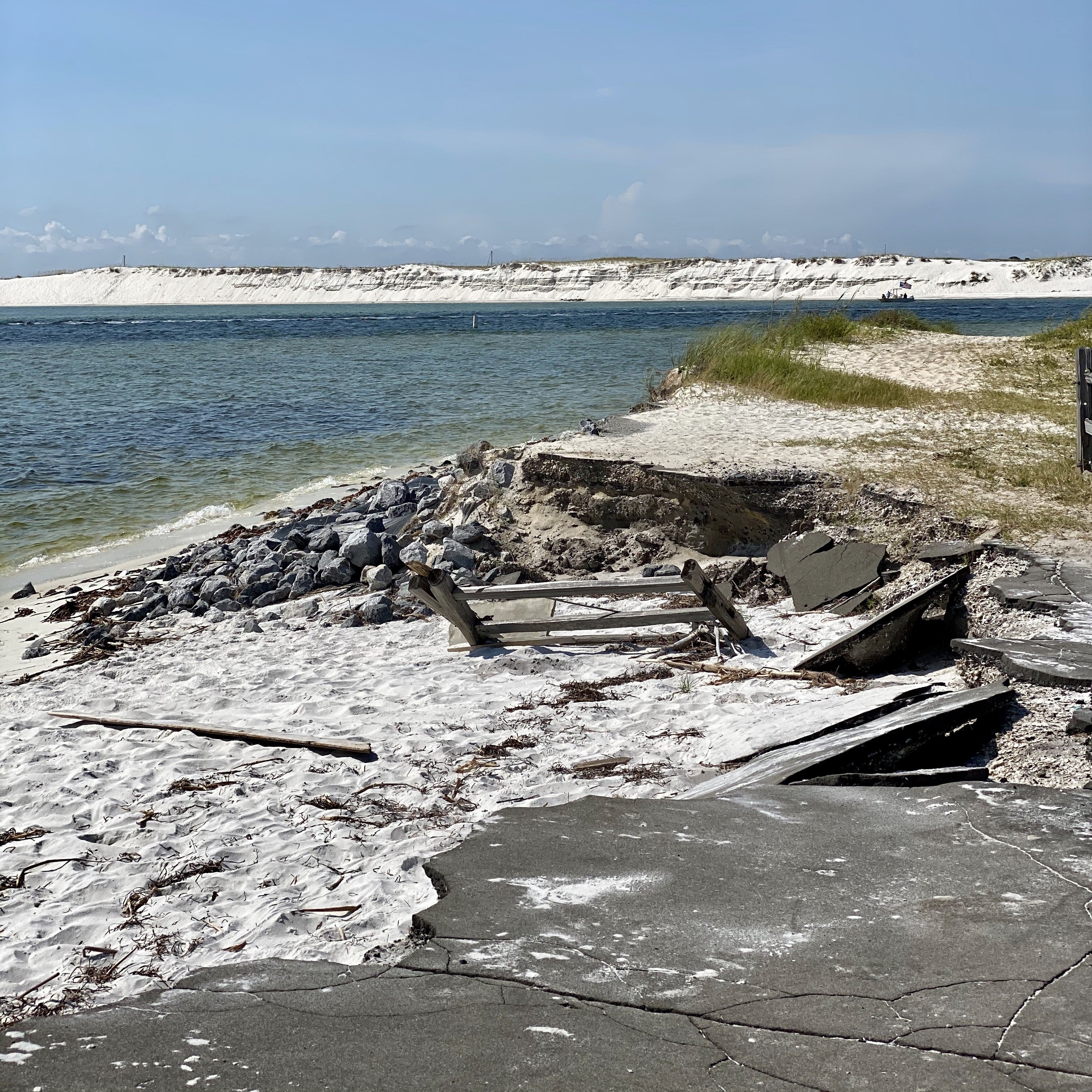 Crumbling asphalt near a sandy beach and blue water, with tall white sand dunes in the distance.
